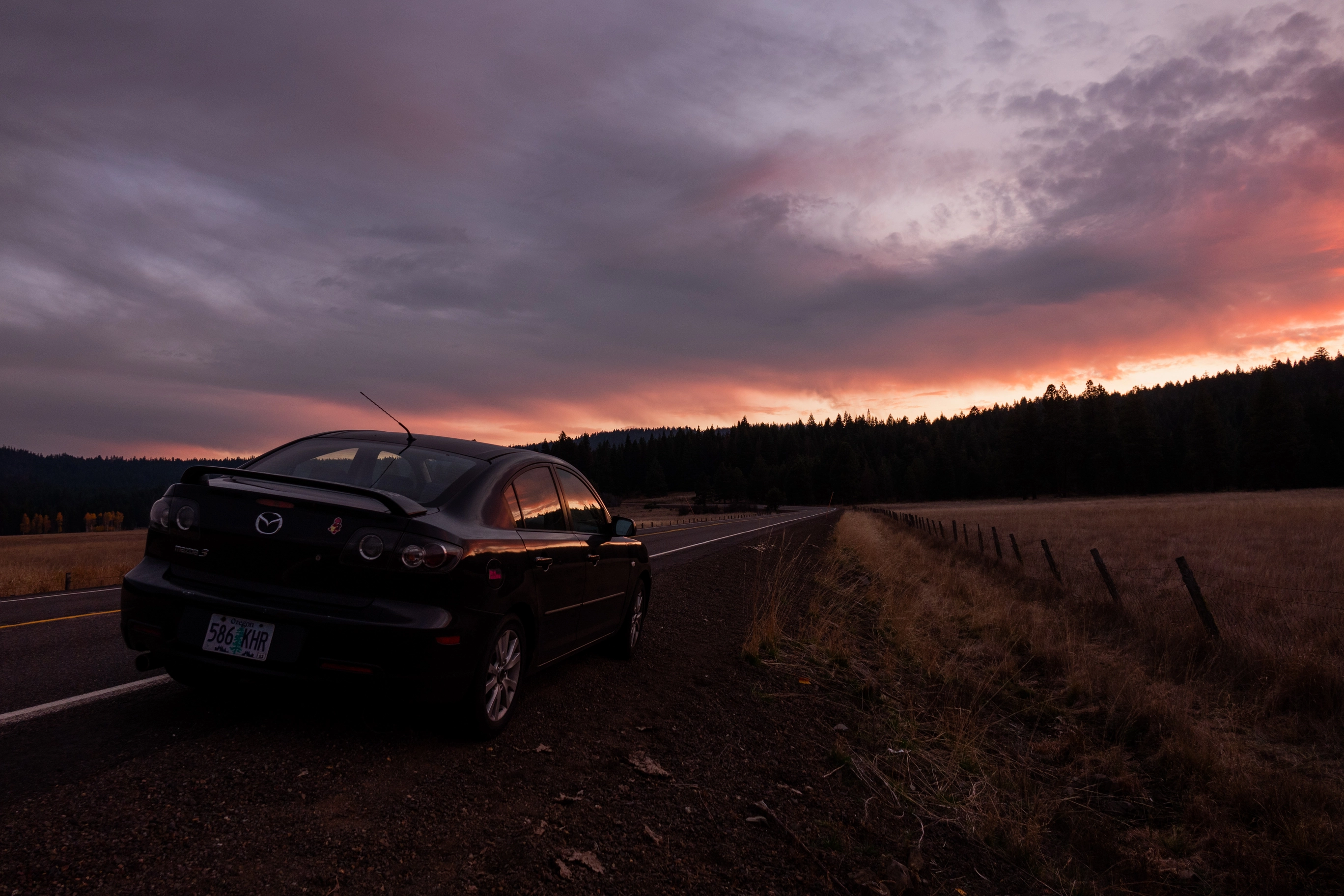 A photo of a black Mazda on the side of a country road during sunset
