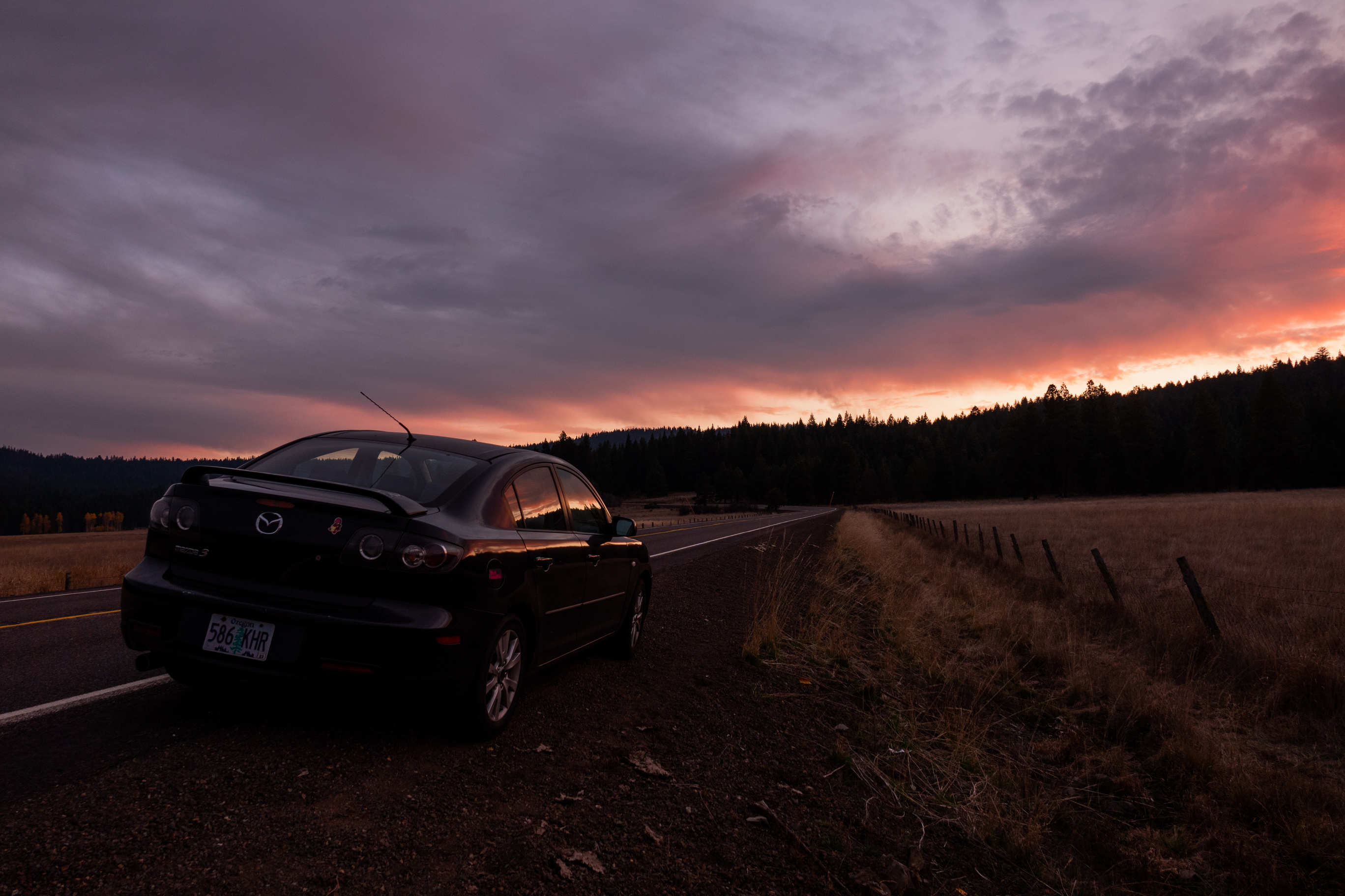 A photo of a black Mazda on the side of a country road during sunset