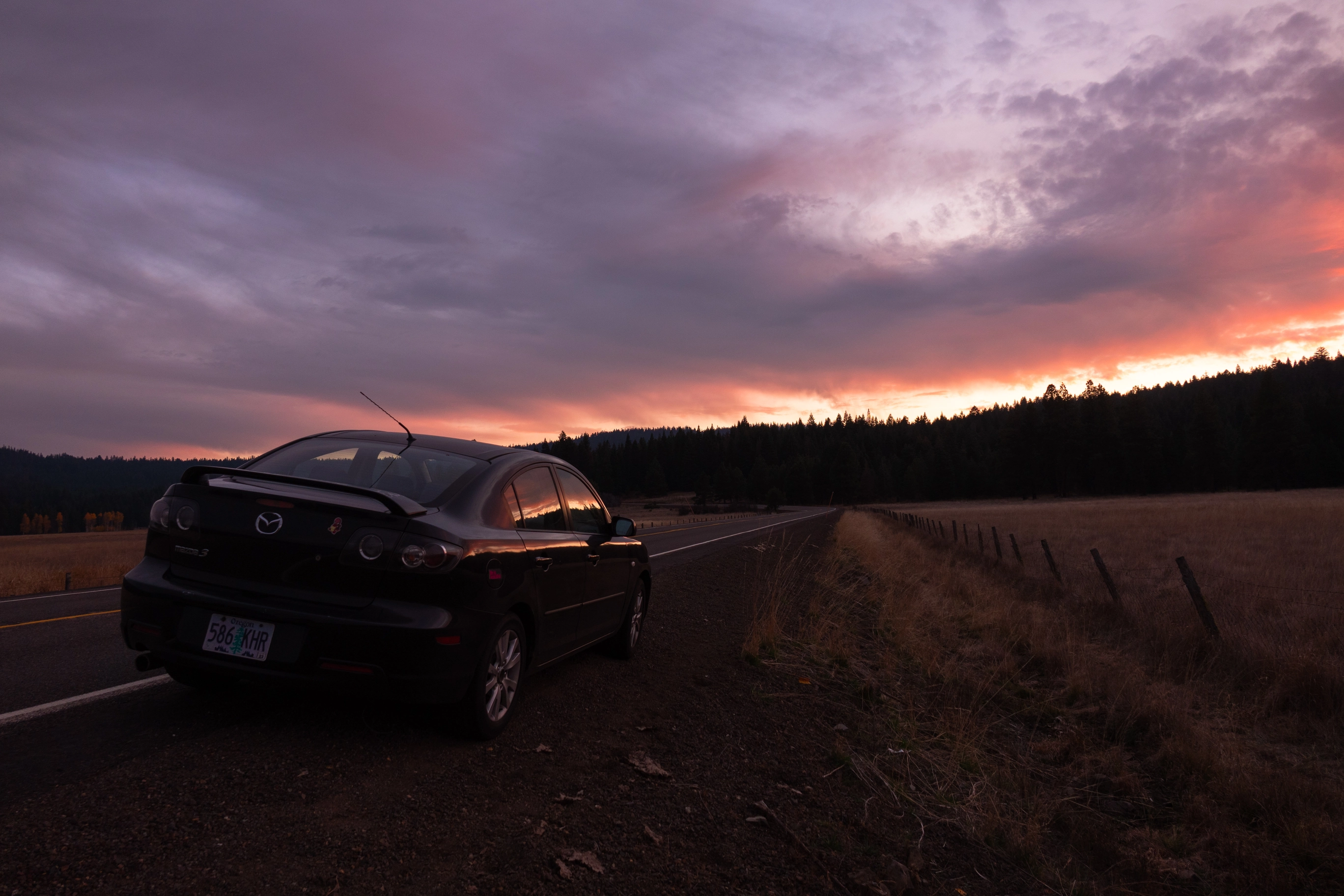 A photo of a black Mazda on the side of a country road during sunset